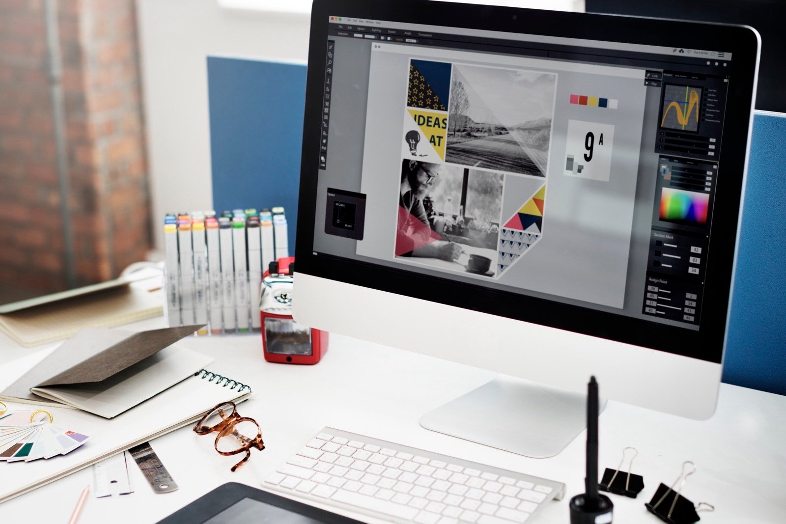 A designer's desk with an iMac showing a layout in progress beside marker pens and colour swatches